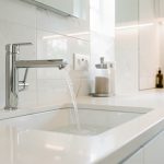Qualité de l’eau maison : quelles solutions selon vos usages quotidiens ? Cuisine et salle de bain modernes, un robinet laisse couler une eau claire, éclairage lumineux et ambiance domestique, surfaces blanches et métal brillants.