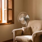 Intérieur apaisant d’un appartement pendant une canicule, avec volets fermés, ventilateur sur pied et thermomètre-hygromètre près d’un fauteuil.