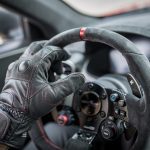 Que signifie la norme FIA 8856-2018 pour les gants de pilote ? Gros plan réaliste sur les gants ignifugés d’un pilote automobile tenant le volant dans l’habitacle d’une voiture de course, avec une ambiance sobre de paddock.