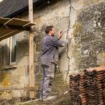 Quels travaux faut-il prioriser pour sécuriser une maison à rénover ? Maison ancienne en rénovation avec échafaudage, tuiles déposées et artisan inspectant les fissures de la façade à la lumière naturelle.