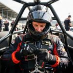 Les équipements de sécurité en compétition automobile : les 8 indispensables Un pilote automobile portant un casque et une combinaison de course, assis dans une voiture de compétition avec un harnais visible dans un paddock.