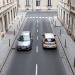 Citadine ou compacte : quelle voiture choisir pour rouler en ville ? Vue réaliste en hauteur d'une citadine et d'une voiture compacte garées dans une rue urbaine européenne étroite, avec des marquages au sol visibles et une lumière naturelle.